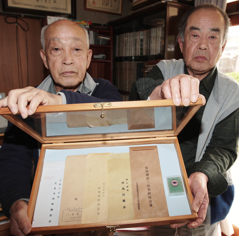封筒に印刷された団体名で顕彰会の変遷を振り返る春野さん(右)と若松さん。ケースの右にあるのは明治４０年制作の発足記念バッジ