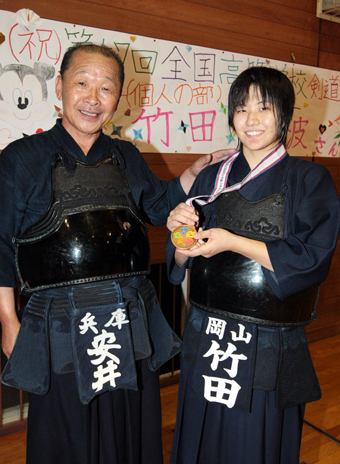 恩師の安井敬明さん(左)にメダルを見せる竹田香波選手。後ろの横断幕は後輩団員の手作り