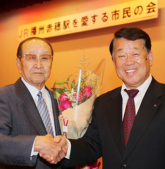 新会長の飯尾義明氏(右)と１０年間の功労に花束を贈られた寒川信男・名誉会長